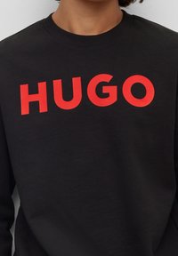 Sudadera negra de algodón con cuello redondo. Presenta un atrevido texto rojo "HUGO" en una fuente moderna en el pecho. Diseño simple de color sólido.