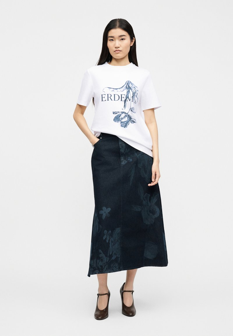 Vrouw draagt een wit T-shirt met bloemenprint, gecombineerd met een donkerblauwe midi-rok met bloemenprint en bruine Mary Jane-hakken, staand met één hand in haar zak.