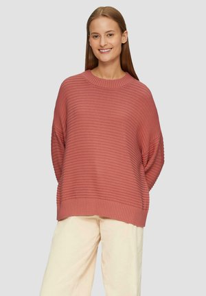 Roze gebreide trui met ribbelstructuur, oversized pasvorm en brede mouwen, gecombineerd met lichtbeige broek.