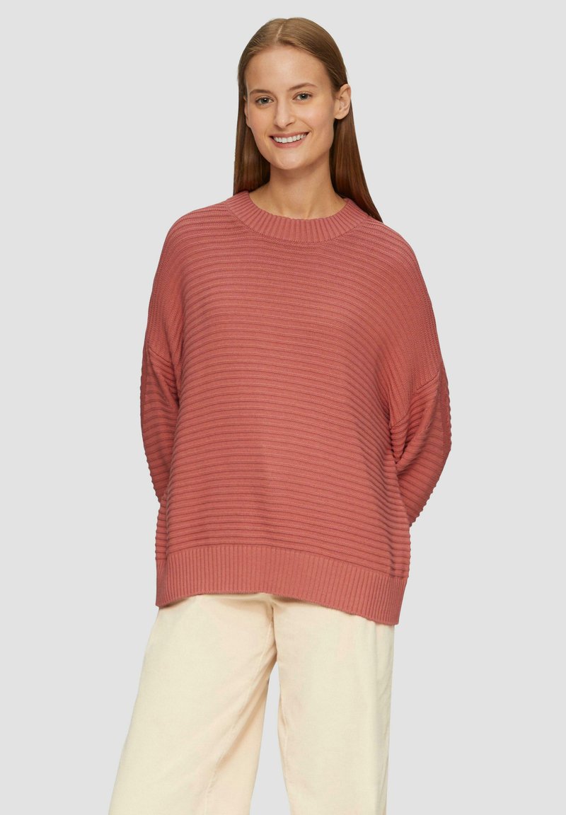 Roze gebreide trui met ribbelstructuur, oversized pasvorm en brede mouwen, gecombineerd met lichtbeige broek.