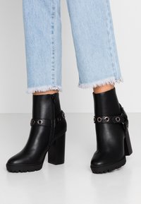 Botas de cuero negro hasta el tobillo con un tacón grueso, que cuentan con una correa con ojales de metal y una cremallera lateral. Suela texturizada para agarre.