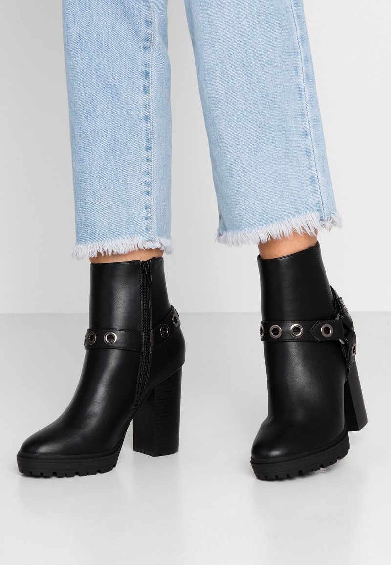 Botas de cuero negro hasta el tobillo con un tacón grueso, que cuentan con una correa con ojales de metal y una cremallera lateral. Suela texturizada para agarre.
