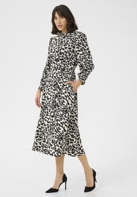Leopardprint jurk van zachte stof, met een knoopsluiting aan de voorkant, een cinched taille, lange mouwen en zijzakken. Zwarte hoge hakken gedragen.
