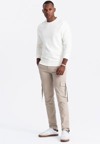 Bílá mikina s žebrovanými manžetami, béžové cargo kalhoty s bočními kapsami a bílé tenisky s hnědými podrážkami. Jednoduchý, volný střih.