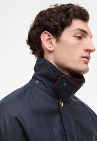 Ναυτικό μπλε σακάκι με ψηλό γιακά, διακοσμημένο με κόκκινη ραφή και χρυσά αξεσουάρ. Κατασκευασμένο από απαλό, αδιάβροχο ύφασμα με προσαρμοσμένη εφαρμογή.