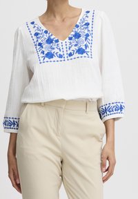 Camicia bianca con ricami floreali blu sul collo e sulle maniche, con design a scollo a V e tessuto testurizzato. Pantaloni beige.