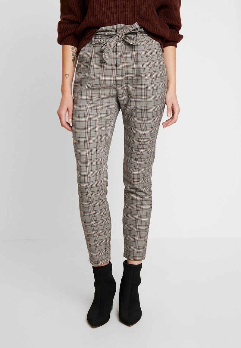 Femme portant un pantalon à carreaux gris taille haute avec une ceinture en tissu nouée à la taille, assorti d'un pull marron et de bottines noires.