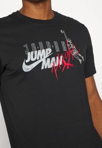 Mustat puuvillapaitat, jossa on graafinen printti, jossa lukee "JORDAN JUMP MAN" hopeisella ja punaisella sekä koripalloilijan siluetti. Pehmeä tekstuuri.