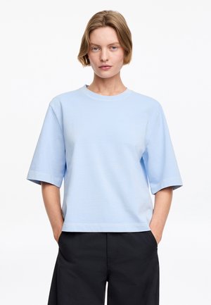 Femme aux cheveux blonds courts portant une chemise bleu clair surdimensionnée à manches courtes et un pantalon noir, debout les mains dans les poches.