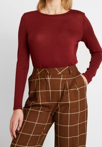 Burgundy långärmad topp i kombination med bruna rutiga höga byxor med ett rutig mönster och sidofickor. Slät textur, åtsittande design.