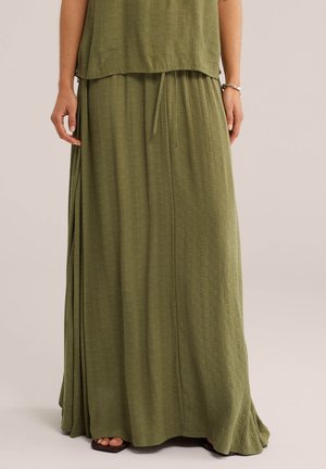 Kobieta ubrana w oliwkową, teksturowaną, długą spódnicę z troczkiem w talii i dopasowaną górę, stoi z rękami swobodnie opartymi wzdłuż boków.