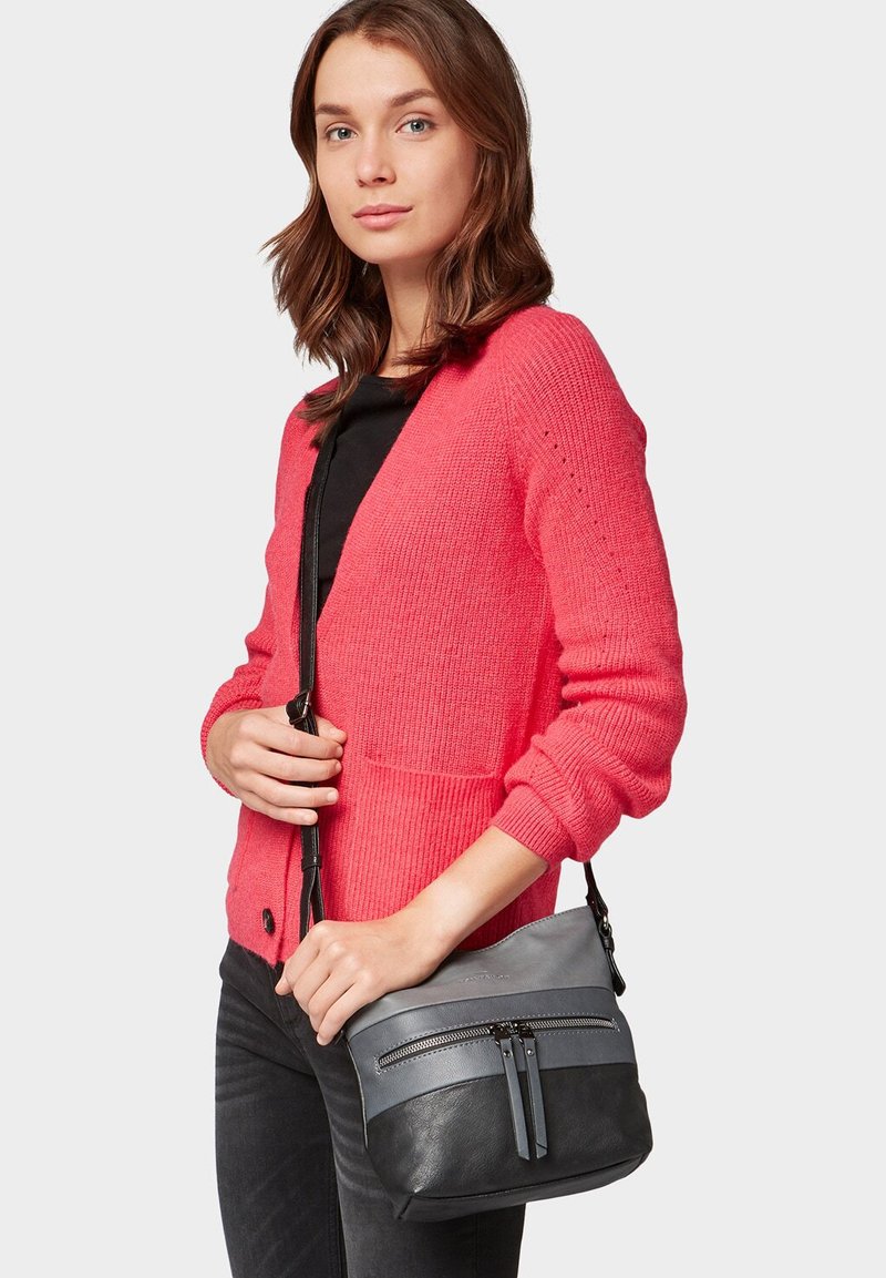 Sac bandoulière en cuir lisse et sombre avec des accents gris, dotée de deux poches zippées et d'une forme structurée. Porté avec un cardigan rose.