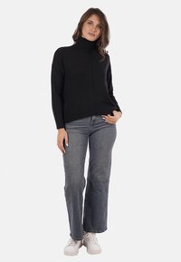 Donna che indossa un maglione nero a collo alto, jeans grigi e sneakers bianche, in piedi con una mano appoggiata sulla vita.