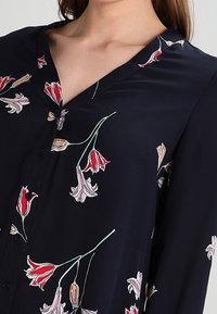 Blusa floreale navy in tessuto leggero, con motivi di tulipani rossi, rosa e bianchi, scollo a V e chiusura con bottoni. Texture liscia.