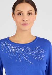 Suéter de punto azul con un escote redondeado y adornos decorativos en varios colores que crean un patrón de olas en la parte superior izquierda.