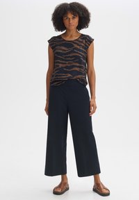 Schwarzes Oberteil mit bronzefarbenem Tigerstreifenmuster, kurze Ärmel, kombiniert mit weiten, marineblauen Hosen und braunen Sandalen mit Plateau-Sohle.