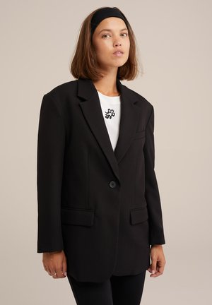 Černý Blazer se strukturovanými rameny, zapínáním na jeden knoflík a bočními kapsami, přeložený přes bílou košili s černým abstraktním vzorem.