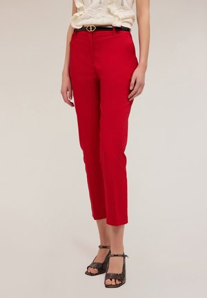 Vrouw met rode cropped broek met zwarte riem en open hakken met slangenhuidmotief, gecombineerd met een wit mouwloos topje.