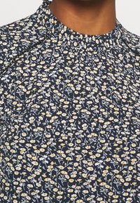 Primer plano de una blusa de cuello alto en un oscuro azul marino con un pequeño patrón floral en blanco y amarillo, llevada por una persona con piel de tono marrón medio.