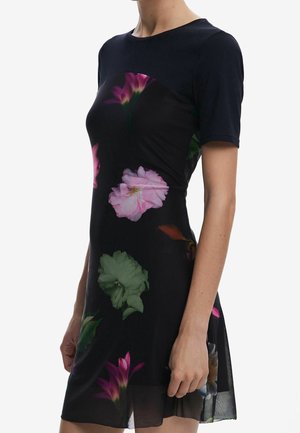 Frau trägt ein kurzärmeliges schwarzes Kleid mit pinken, grünen und orangefarbenen Blumenmustern und steht vor einem einfachen weißen Hintergrund.