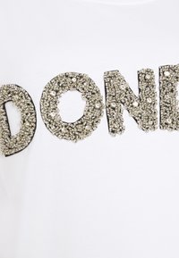 White t-shirt featuring the word "DONE" decorated with silver sequins and beads, outlined in black, creating a textured, dimensional effect.