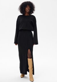 Czarna dzianinowa sukienka z długimi rękawami i rozcięciem do uda, w zestawieniu z beżowymi kozakiem za kolano. Materiał ma delikatny połysk.