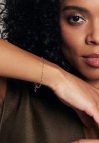 Złota bransoletka na nadgarstku, z owalnymi ogniwami i zapięciem typu homar. Kolaż skóry ma średni odcień, a w tle znajduje się teksturowana brązowa tkanina.