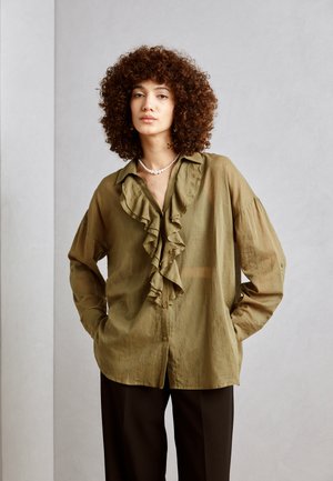 Blusa verde oliva, transparente, de manga larga con un pliegue en la parte delantera, cierre de botones y ajuste holgado, combinada con pantalones negros.