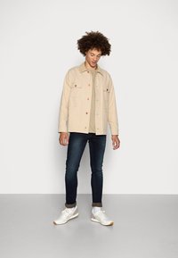 Beige bomullsjakke med knappelukking og to frontlommer, båret over en lysegrå skjorte og mørke jeans, kombinert med hvite joggesko.