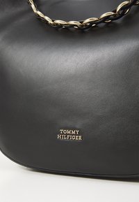 Borsa a mano in pelle nera con una texture liscia, dotata di un manico a catena dorata e di una placca con logo dorato che reca la scritta "TOMMY HILFIGER."