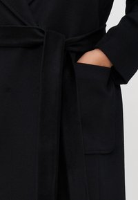 Cappotto di lana nero con vita cintrata, ampio tascone e maniche larghe. Il materiale ha una texture liscia e un design su misura.