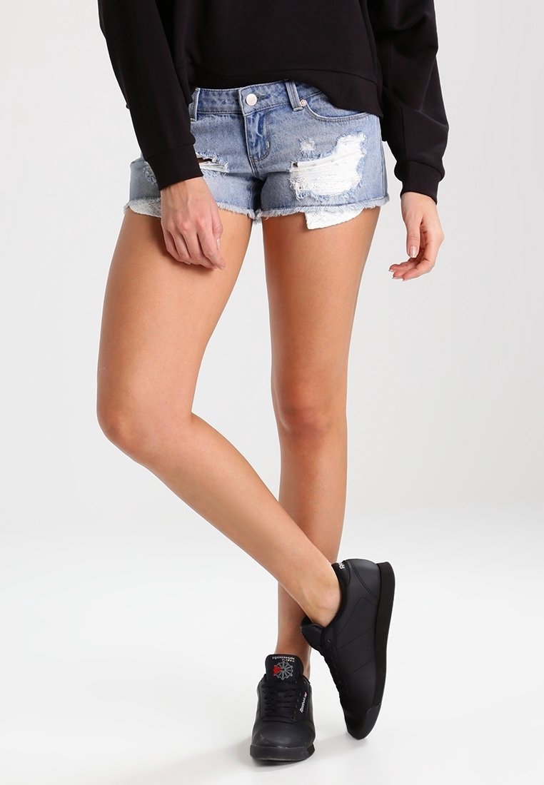 Shorts vaqueros de color azul claro con detalles desgastados, dobladillo deshilachado y diseño de cinco bolsillos, combinados con zapatillas negras.