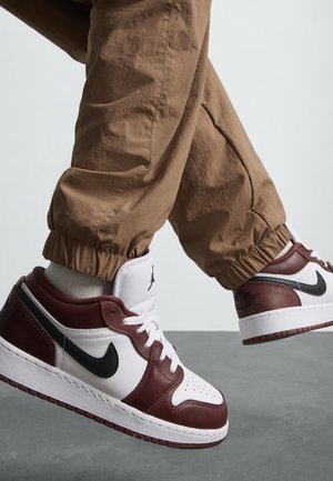 Baskets en cuir blanc et bordeaux avec des accents noirs, à bout rond et à lacets. Portées avec un pantalon beige à cordon de serrage, mettant en valeur des bas de pantalon retroussés.
