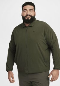 Alyvmedžio žalia Nike striukė su užtrauktuku, apykaklės dizainu, ilgomis rankovėmis su elastingais mėgintuvėliais ir mažas juodas logotipas ant krūtinės.