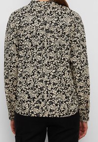 Femme portant un chemisier à manches longues noir et beige avec un motif floral dense, vue de dos, assorti à un pantalon noir.