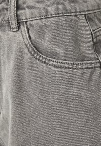 Gros plan sur un tissu en denim gris montrant la zone de la poche avant avec des coutures détaillées et une boucle de ceinture sur un jean.