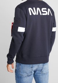 Sweatshirt bleu marine avec le logo "NASA" blanc au dos, des rayures blanches sur les manches et une poche zippée sur le bras gauche.