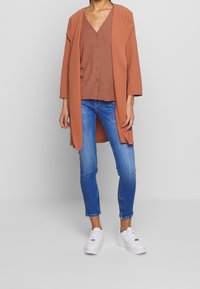 Cardigan lungo color ruggine sopra un top a V color ruggine, abbinato a jeans skinny blu e sneakers bianche. Il cardigan presenta un design con apertura frontale.