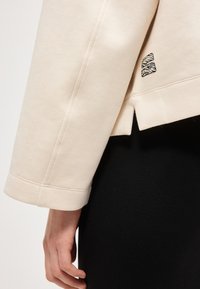 Haut beige à manches longues avec fente latérale et petit logo sportif noir et blanc sur la hanche, porté avec un pantalon noir, main détendue le long du corps.