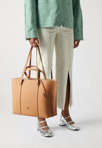 Brahna ādas somi ar strukturētu formu, divām augšējām rokturiem un iepresētu logo. Valkāta ar piparmētru zaļu žaketi un krēmkrāsas biksēm.