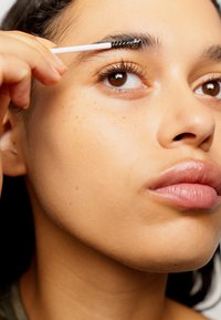Nahaufnahme einer Person, die ihre Augenbrauen mit einer schwarzen Wimpernbürste pflegt. Die Haut wirkt glatt mit einem natürlichen, gleichmäßigen Hautton. Braune Augen sind sichtbar.