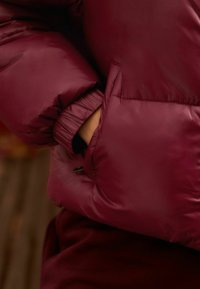 Bordeaux pufferjack met elastische manchetten en een zijzak. Glad, glanzend materiaal met een geborduurde textuur, ontworpen voor warmte en comfort.