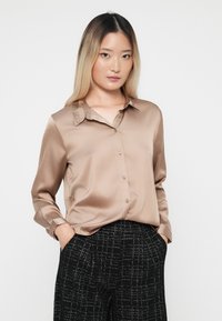 Camicia button-up beige in satin con colletto, maniche lunghe e una texture liscia, abbinata a pantaloni a scacchi bianchi e neri.