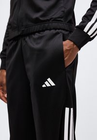 Άτομο που φοράει μαύρη φόρμα Adidas με τρεις λευκές ρίγες στο πλάι και το χέρι στηρίζεται στην τσέπη, με απλό φόντο.