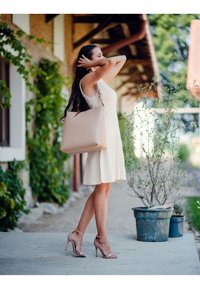 Große, weiche rosa Tragetasche mit minimalistischem Design und ohne sichtbare Beschläge oder Muster, gehalten von einer Person in einem hellen cremefarbenen Kleid.