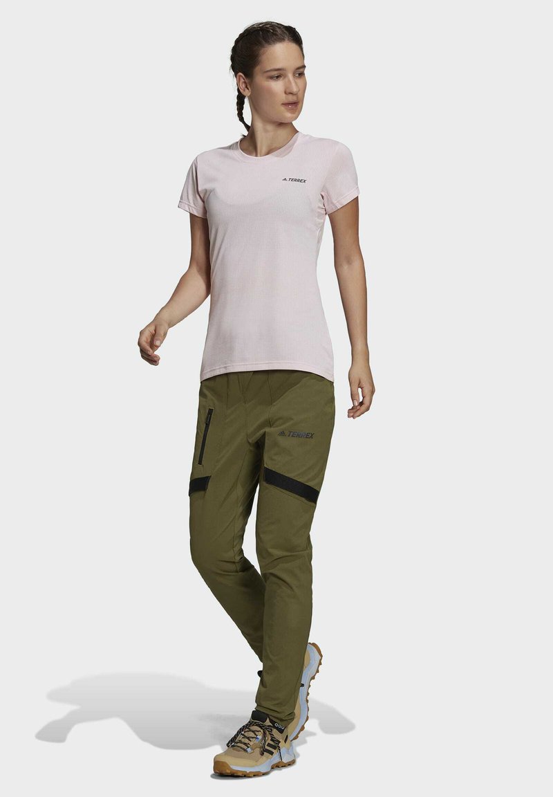 Helles rosa Kurzarmhemd mit Logo; olivgrüne Hose mit Reißverschlüssen und schwarzen Klettakzenten; beige Wanderschuhe mit dunklen Schnürsenkeln.