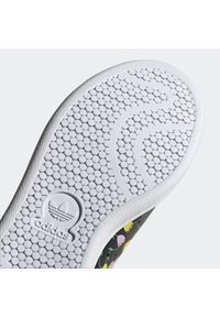 Bílá podrážka boty Adidas s kruhovým vzorem protiskluzu a logem Adidas, na světlém pozadí je viditelná část nártu boty s květinovým vzorem.