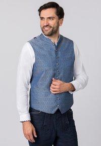 Blaues Weste mit komplexem Paisleymuster, metallischen Knöpfen und Stehkragen, getragen über einem weißen Hemd und dunklen Hosen.