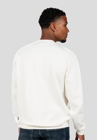 Felpa leggera color crema con collo a giro, texture morbida e polsini a costine. Presenta una cucitura centrale sulla schiena e un logo discreto sul collo.