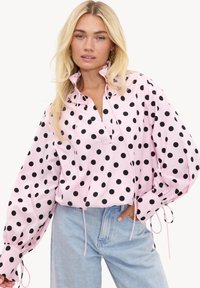 Femme blonde portant un chemisier rose clair avec des pois noirs, col à volants, manches bouffantes et jean bleu clair, posant avec une main dans la poche.